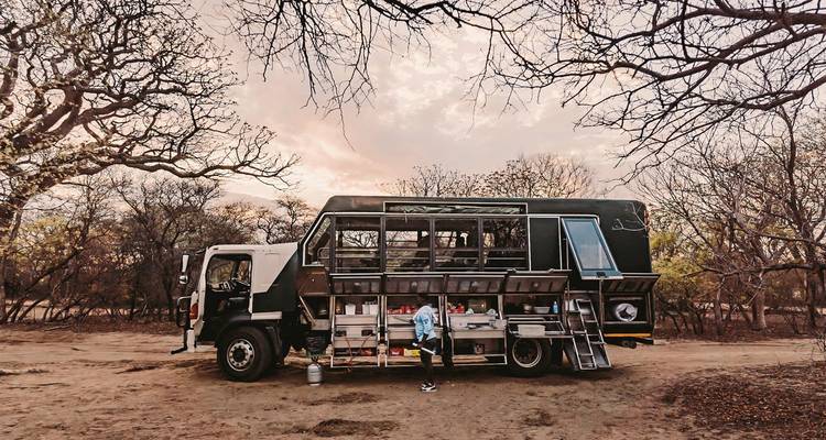Un camión de safari con compartimentos abiertos estacionado en un área seca y boscosa.