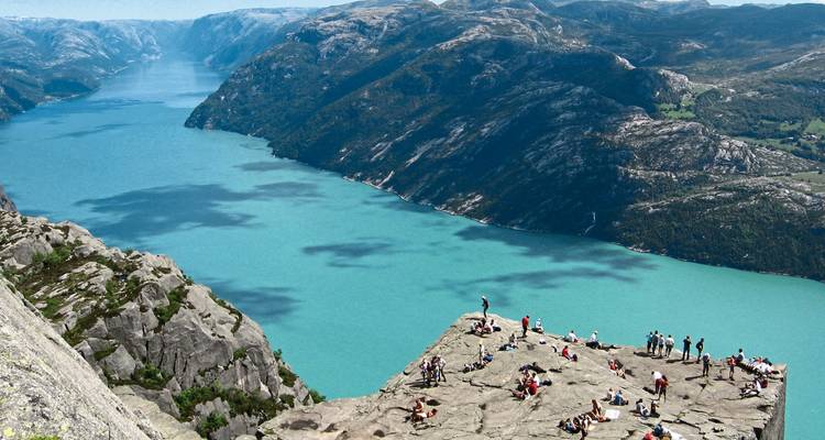 Des touristes debout sur une falaise surplombant un fjord bleu serpentant entre les montagnes.