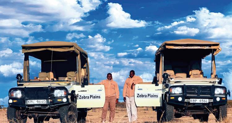 Dos personas paradas junto a vehículos de safari bajo un cielo dramático.