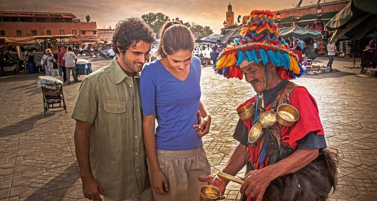 A man in traditional attire showing items to a couple in a market square.