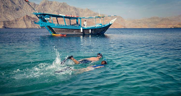 Twee mensen die snorkelen bij een traditionele boot tegen een bergachtige achtergrond.