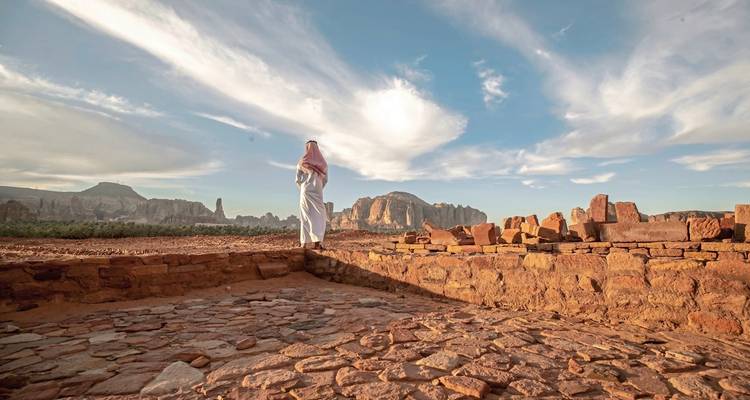 Person in traditional attire standing amidst ancient ruins in the desert.