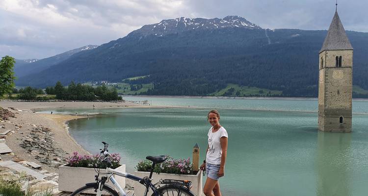 Mujer junto a torre de iglesia sumergida en lago con montañas.