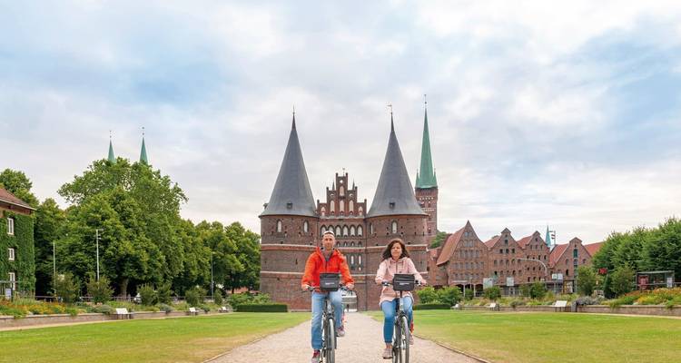 Ciclistas pedaleando hacia una puerta histórica con múltiples torres.