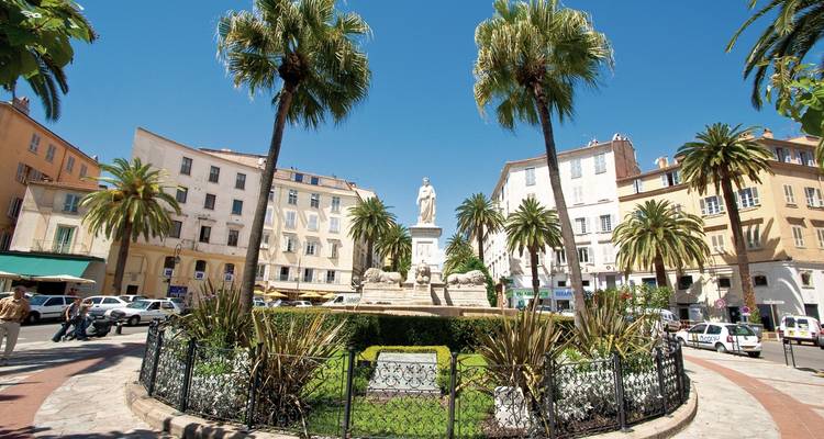 Een stadsplein met een standbeeld omringd door palmbomen en klassieke gebouwen.