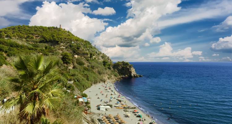 Malerischer Strand mit Menschen, die sich unter Sonnenschirmen entspannen, umgeben von Grün und einem klaren blauen Meer.