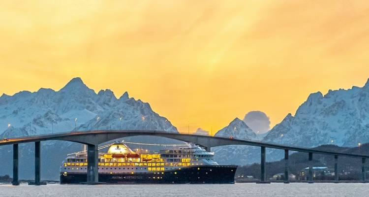 Ein großes Kreuzfahrtschiff, das bei Sonnenuntergang unter einer Brücke mit bergiger Landschaft hindurchfährt.