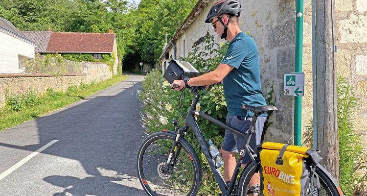 Cyclist checking a map on a country road flanked by greenery and quaint buildings.