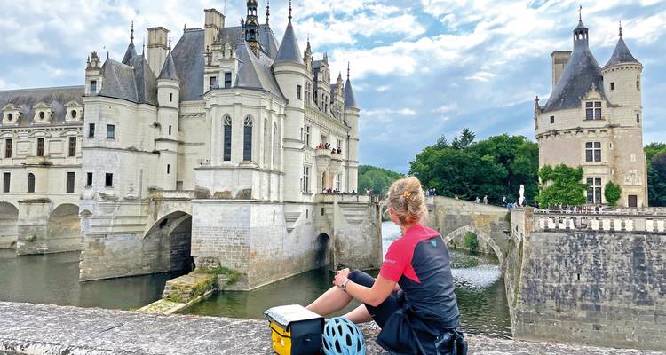Cyclist sitting beside a river, observing a grand chateau with turrets.