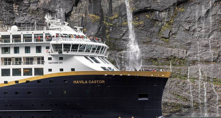 A close-up view of a cruise ship near a waterfall in a fjord.