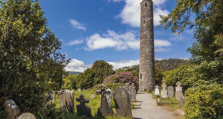 Runder Steinturm, der sich über einen alten Friedhof erhebt, umgeben von grünen Bäumen unter einem blauen Himmel mit zerstreuten Wolken.
