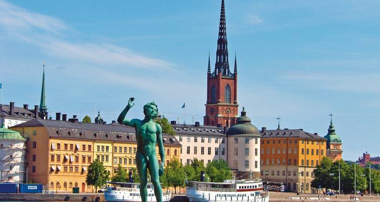 Statue near colorful buildings, waterfront, and spire.