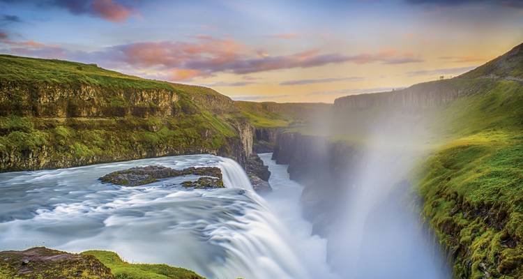 Kraftvoller Wasserfall in einer grünen Landschaft unter einem farbenfrohen Himmel.