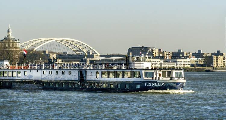 Cruiseschip varend op een rivier met moderne stad op de achtergrond.