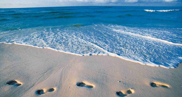 Fußspuren im feinen Sand neben sanften Wellen an einem blauen tropischen Meer.