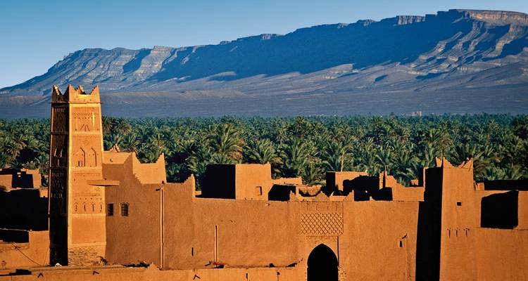 Lehmziegelgebäude umgeben von Palmen mit Bergen im Hintergrund.