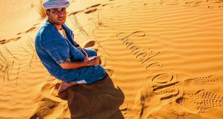 Eine Person in traditioneller Kleidung, die auf Wüstensand sitzt, mit 'Sahara' daneben geschrieben.