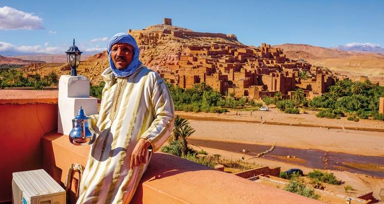 Eine Person in traditioneller Kleidung, die eine Teekanne hält, mit Ait Benhaddou im Hintergrund.