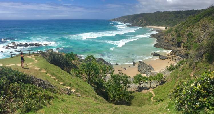A panoramic view of a coastal landscape with a person standing near the shore, lush greenery, and waves crashing against the rocks.