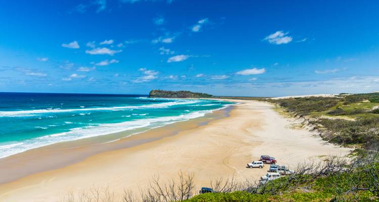 Une longue plage de sable avec des vagues turquoise et quelques véhicules garés sur le sable.