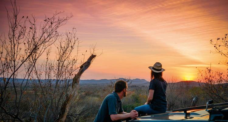 Deux personnes assises sur le toit d'une voiture regardant le coucher de soleil sur la savane.
