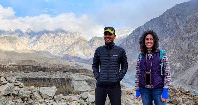 Two trekkers standing on rocky terrain with dramatic mountains shrouded in cloud behind them.