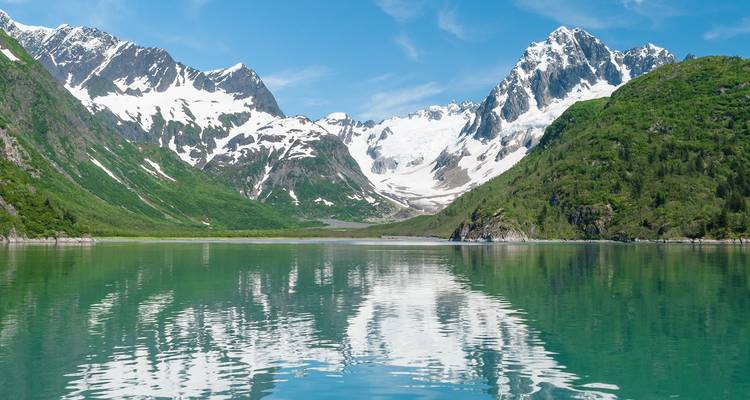Un lago glacial de color turquesa que refleja perfectamente los picos nevados y las laderas verdes bajo un cielo azul despejado.