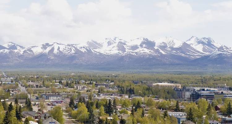 Vista aérea de una ciudad rodeada por una vasta cordillera cubierta de nieve y bosque verde en primavera.