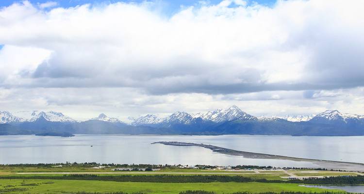 Escena costera panorámica con una lengua de tierra larga y estrecha que se extiende hacia aguas tranquilas, picos nevados a lo lejos y campos verdes en primer plano bajo cielos parcialmente nublados.
