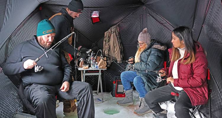Des amis pratiquant la pêche blanche à l'intérieur d'une cabane isolée sombre, assis autour d'un trou percé avec des cannes à pêche et de l'équipement sur un lac gelé.