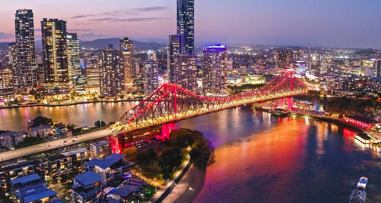 El Puente Story Bridge de Brisbane iluminado en un rojo vivido que se extiende sobre el río al atardecer con rascacielos más allá.