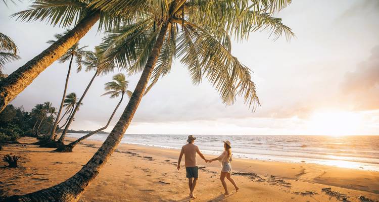 Una pareja que se da la mano camina por una playa tropical bordeada de palmeras al atardecer.