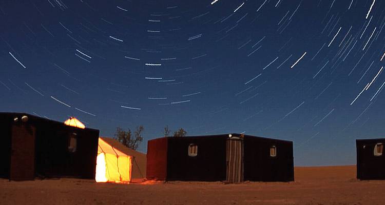 Sahara desert camp beneath star-trail night sky with glowing tent entrance and dark sand.