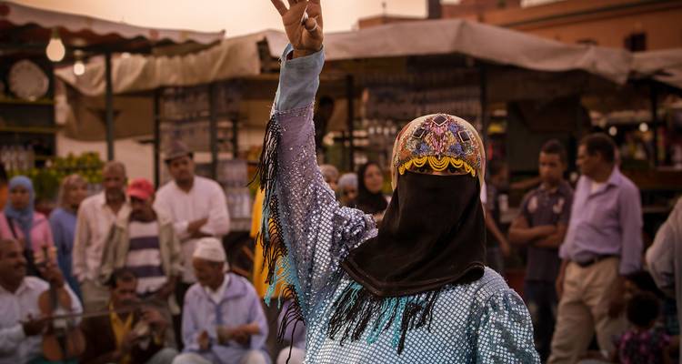 Street performer in sequined costume raises a peace sign while an audience of locals watches in a Marrakesh market at dusk.