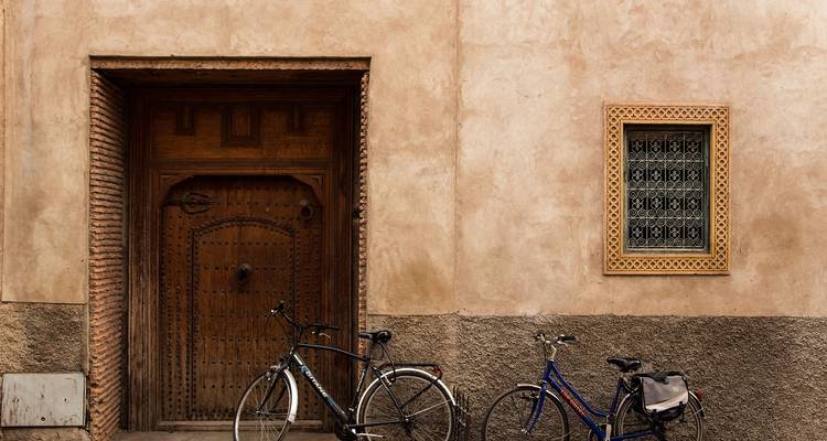 Weathered wooden door and small ornate window set in a clay-coloured wall with two bicycles leaning casually outside.