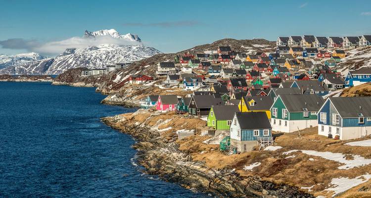 Des maisons en bois colorées perchées sur une côte rocheuse avec des montagnes enneigées en arrière-plan au Groenland.