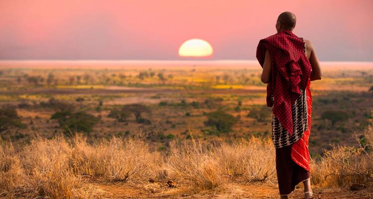 Maasai-Mann in traditioneller Shuka mit Blick auf die weite Savanne während die orange Sonne am Horizont untergeht.