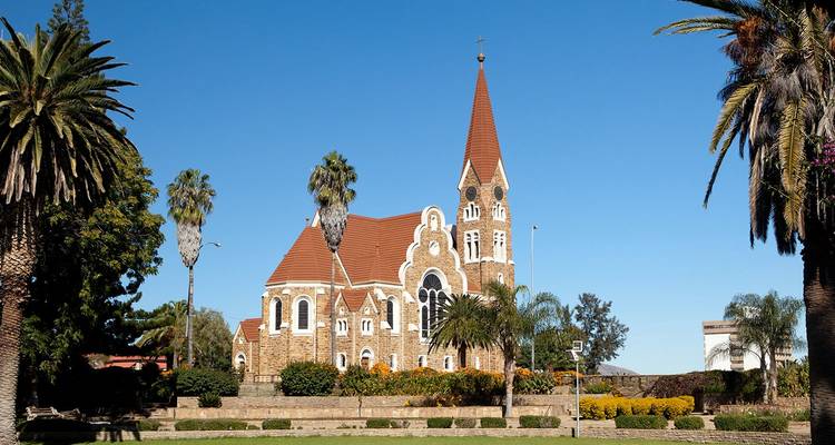 La iglesia Christuskirche con techo rojo en Windhoek se alza enmarcada por palmeras contra un cielo despejado.
