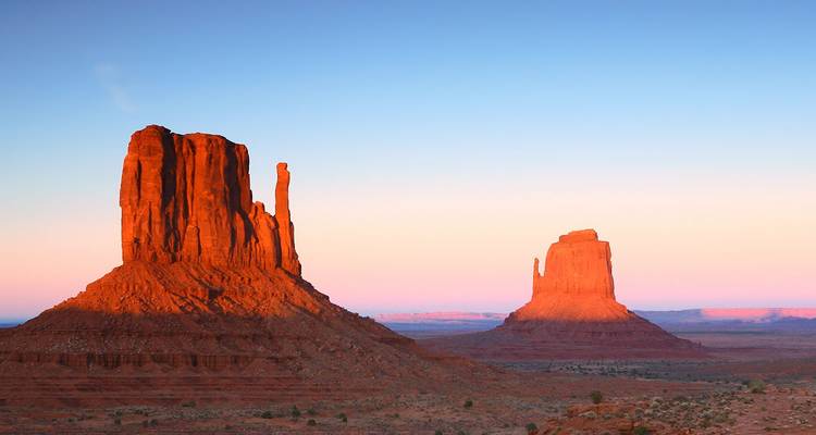 Twin buttes of Monument Valley glowing orange in early morning light over the desert floor