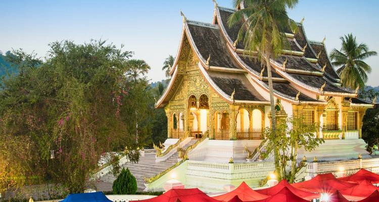 Der beleuchtete Tempel Haw Pha Bang leuchtet hinter farbenfrohen Marktschirmen des Nachtmarkts in Luang Prabang.
