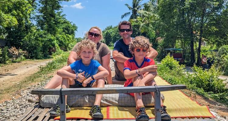 Smiling family of four seated on a bamboo train cart riding through lush countryside.