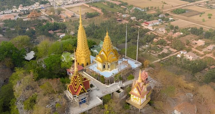 Aerial view of hilltop temples with golden spires surrounded by Cambodian countryside.
