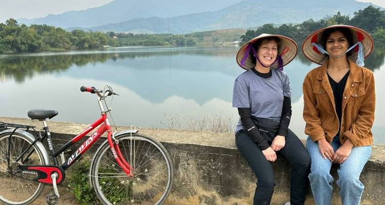 Two travellers in conical hats relaxing by a tranquil lake with mountains beyond and a red bicycle.