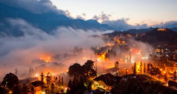 Nebelige Stadtlandschaft in der Dämmerung mit beleuchteten Straßen und Bergkulisse.