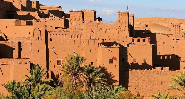 Sunlit ancient kasbah with palm trees in front.