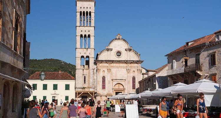 Plaza de piedra concurrida en Hvar con cafeterías al aire libre, sombrillas blancas y la alta torre de campanas de la Catedral de San Esteban.