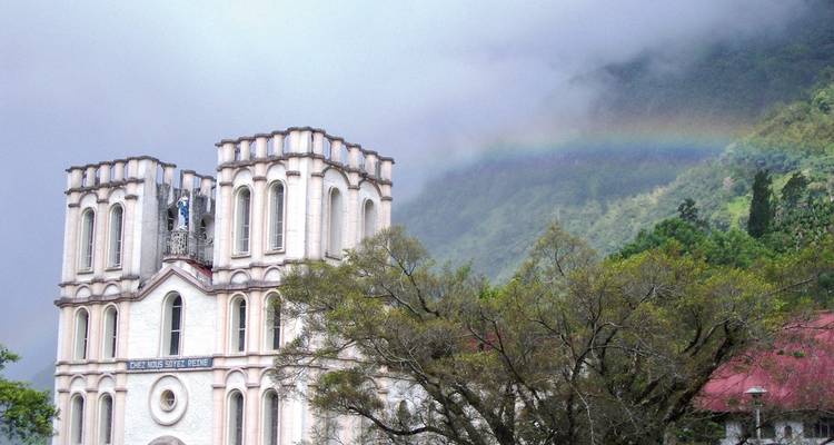 A white colonial church stands at the base of misty green mountains with a vivid rainbow above.