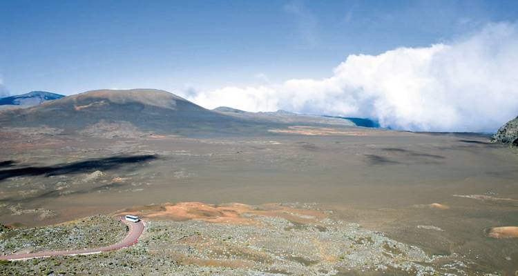A vast volcanic plain stretches to distant slopes beneath a blue sky and low bank of clouds.