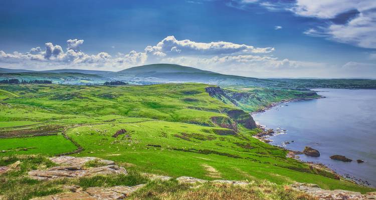 Weelderige groene heuvels aan de kust rollen af naar een kalme blauwe zee onder een heldere lucht in Noord-Ierland.