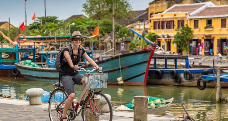 Un cycliste souriant passe devant des bateaux colorés et des maisons-boutiques jaunes le long de la rive fluviale de Hoi An.